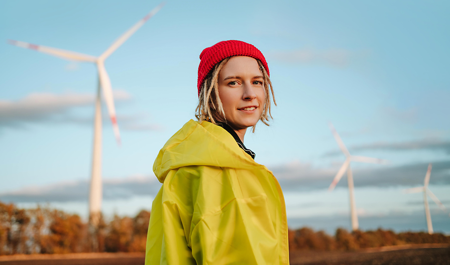 Junge Frau in gelber Jacke und roter Mütze steht vor Windrädern und symbolisiert die gemeinsame Verwirklichung der Energiewende durch die Energiegemeinschaft.